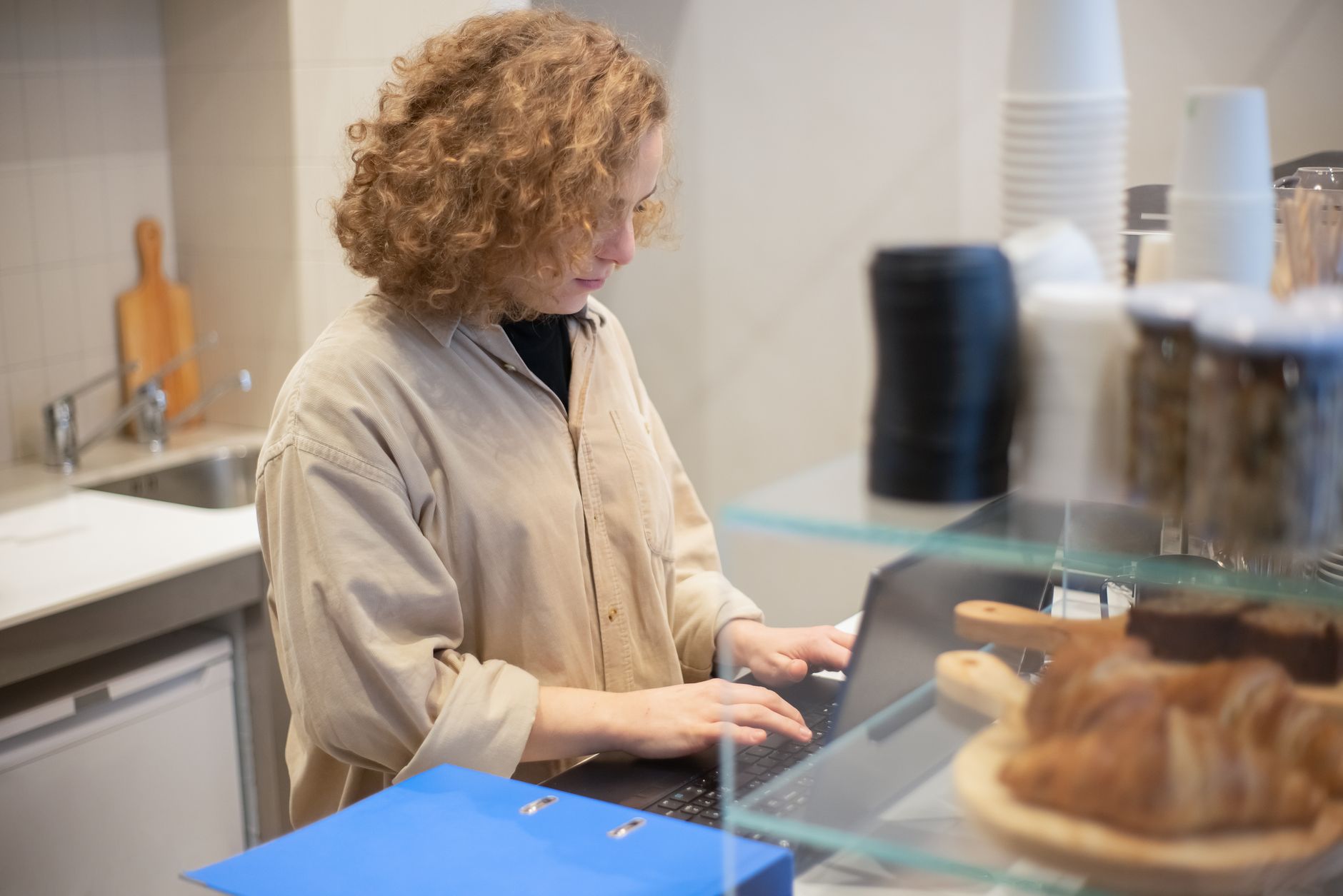 A young woman uses a laptop behind a café counter with pastries in foreground.
